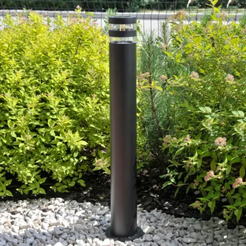 Lampa ogrodowa słupek RINO o wysokości 74 cm, okrągła, czarna. Ustawiona pionowo na tle zielonych krzewów, z przezroczystym kloszem i metalową podstawą, tworzy nowoczesny element oświetlenia zewnętrznego.