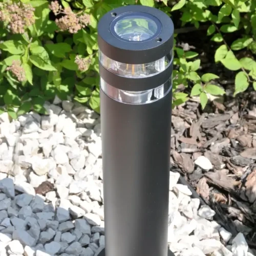 Lampa ogrodowa słupek RINO o wysokości 74 cm, w kolorze czarnym, okrągła, z przezroczystą górną częścią. Ujęcie z boku, na tle zieleni i kamieni. Widoczne detale konstrukcji oraz miejsce na żarówkę E27.