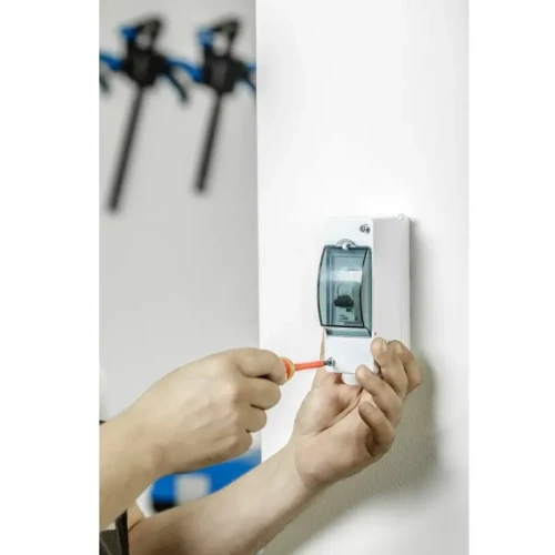 Izolowany wkrętak w kolorze czerwonym, używany do montażu w obudowie elektrycznej na ścianie. W tle widoczne narzędzia montażowe. Wkrętak ma stalowy uchwyt i jest przystosowany do napięcia 1000V.