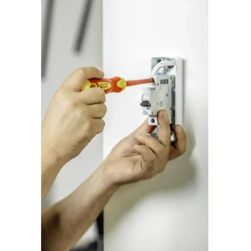Komplet wkrętaków izolowanych 2 szt. 1000V, stal. Wkrętak z czerwoną rękojeścią w użyciu, montujący elementy elektryczne na białej ścianie. Widać detale narzędzia oraz dłonie użytkownika.