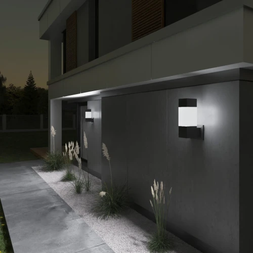 Oprawa architektoniczna INVO E27 w kolorze grafitowym, zamontowana na zewnętrznej ścianie budynku. Prosty, nowoczesny design z białym kloszem. W tle widoczne rośliny ozdobne oraz jasny chodnik. Oświetlenie w nocy, tworzy przytulną atmosferę.