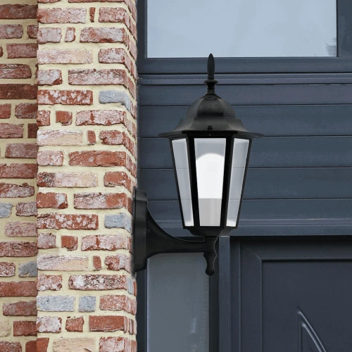Lampa wisząca ogrodowa LIGURIA E27 w kolorze czarnym, zamontowana na ścianie. Posiada szklane panele z przodu, klasyczny kształt, dekoracyjny uchwyt. Umieszczona obok ceglanej ściany, w tle ciemnoniebieskie drzwi.