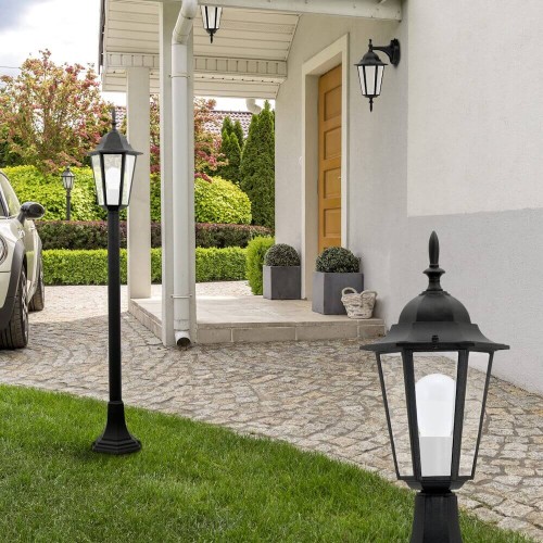 Lampa ogrodowa słupek LIGURIA o wysokości 96,5 cm, w kolorze czarnym, z metalową podstawą i kloszem w kształcie latarni. Ustawiona na trawniku przed domem, obok podjazdu, w otoczeniu roślinności oraz innych lamp ogrodowych.