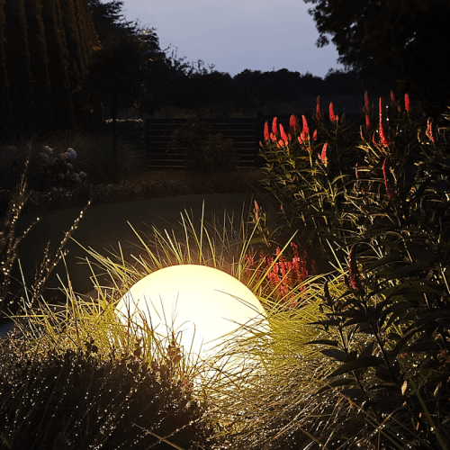 Lampa ogrodowa Kula Globo o średnicy 30 cm, z białym, okrągłym kloszem, umieszczona wśród wysokiej trawy i kolorowych kwiatów, w otoczeniu zieleni, podświetlająca przestrzeń w ciemności. Ujęcie z boku, z bliska.