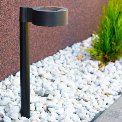 Lampa solarna EYE IP44 0,06W w kolorze czarnym o wysokości 38 cm, umieszczona na tle żwirem i zielonymi roślinami, z górnym panelem słonecznym widocznym na górnej części. Ujęcie frontalne.