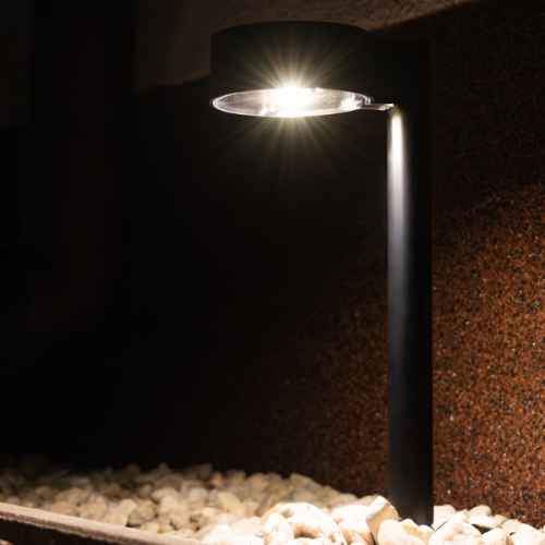 Lampa solarna EYE IP44 0,06W w kolorze czarnym, wysokość 38 cm. Zainstalowana w ogrodzie, z okrągłą głowicą emitującą światło. Kąt ujęcia z boku ukazuje jej elegancki kształt oraz otoczenie z drobnymi kamieniami.