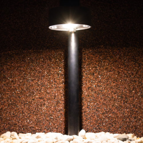 Lampa solarna EYE IP44 0,06W w kolorze czarnym, wysokość 38 cm. Posiada okrągłą głowicę emitującą światło, umieszczoną na prostym, cylindrycznym słupku, otoczona drobnymi kamieniami. Tło o fakturze brązowej.