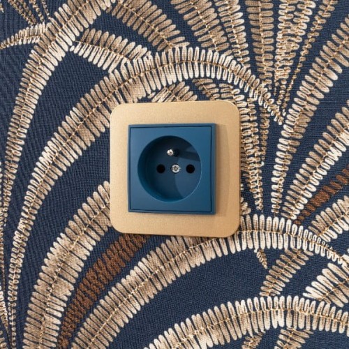Gniazdo podwójne Karlik MINI w kolorze navy blue z uziemieniem, umieszczone w złotej ramce. Widoczny otwór na dwa bolce oraz uziemienie. Tło z wzorem w odcieniach brązu i złota.