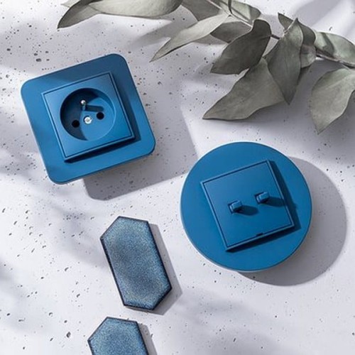 Karlik MINI Ramka pojedyncza w kolorze navy blue, kwadratowa ramka z gniazdkiem i okrągła ramka z włącznikiem. Elementy na jasnym tle, otoczone liśćmi. Widoczne różne kształty oraz faktura materiału.