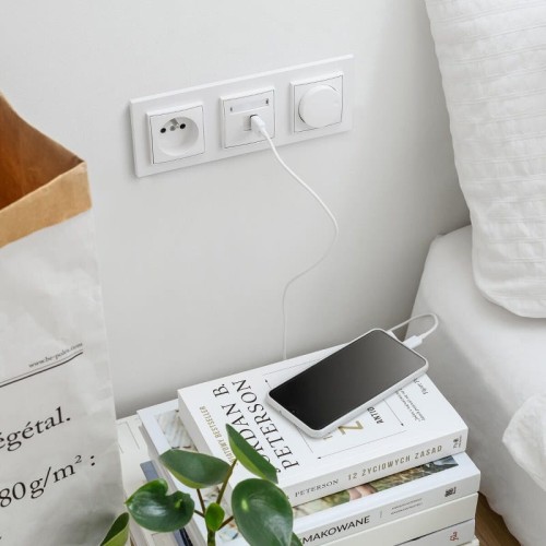 Potrójna ramka Karlik MINI w kolorze białym z trzema gniazdami: gniazdo elektryczne, gniazdo USB oraz włącznik. Ujęcie z boku, zamontowana na ścianie obok książek i telefonu ładowanego kablem. Tło jasne, minimalistyczne.