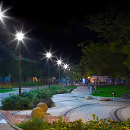 Lampa uliczna RX2 100W 15000LM, zimno biała, umieszczona na słupach oświetleniowych, oświetla nocną scenerię parku z alejkami, z zielonymi krzewami i kamieniami dekoracyjnymi w tle.