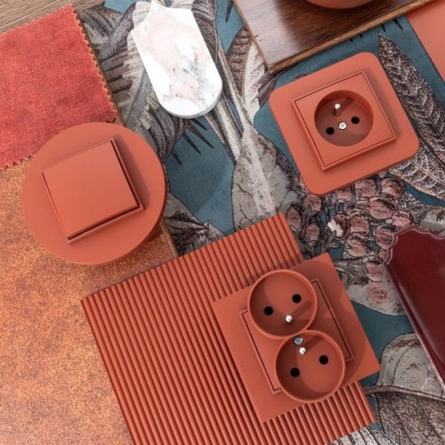 Łącznik podwójny świecznikowy Karlik MINI w kolorze terakota, o kwadratowym kształcie, z widocznymi dwoma gniazdami. Produkt leży na różnorodnych materiałach tekstylnych w odcieniach brązu i zieleni, w stylowej aranżacji.