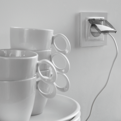 Ramka pojedyncza Simon 10 w kolorze białym, zamontowana na ścianie, z przezroczystą pokrywą. W tle stos białych filiżanek z widocznymi uchwytami i talerzy. Kabel elektryczny wpięty w ramkę, na jasnym tle.