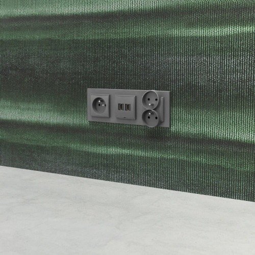 Łącznik Karlik MINI w grafitowym macie, umieszczony na ścianie. Zawiera dwa gniazda elektryczne oraz jedno gniazdo USB. Prosta forma, z widocznymi otworami na wtyczki, na tle ciemnozielonej tekstury ściany.