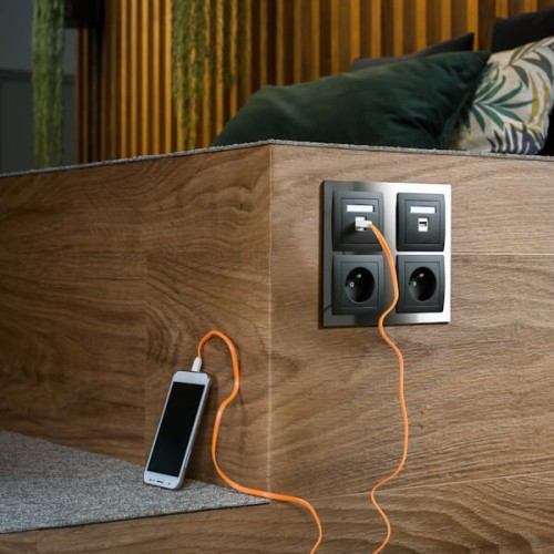 Gniazdo pojedyncze Karlik Deco bez uziemienia w kolorze czarnym matowym, zamontowane na ścianie, z widocznym kablem USB oraz podłączonym telefonem. Tło to drewniana powierzchnia z roślinnymi dekoracjami.
