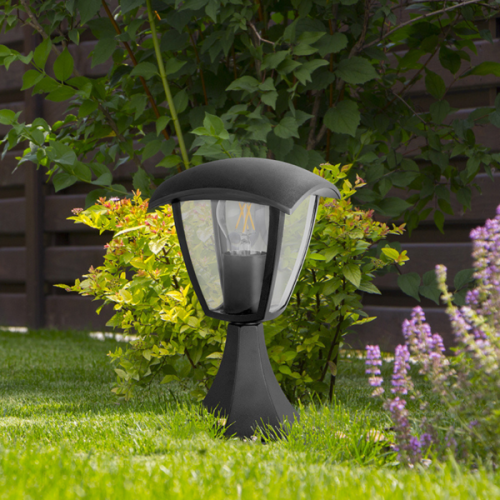 Lampa ogrodowa słupek IGMA, wysokość 29,5 cm, w kolorze czarnym, z przezroczystym kloszem. Produkt umieszczony na trawie, w otoczeniu zieleni i kwiatów, z subtelnym, zaokrąglonym dachem.