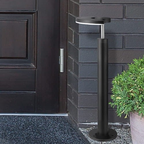 Lampa ogrodowa słupek LED MODENA o wysokości 54 cm w kolorze czarnym. Stoi obok drzwi, ma prostą, cylindryczną formę z metalowym wykończeniem i okrągłą górną częścią, która emituje światło. W tle widoczna roślina w doniczce.