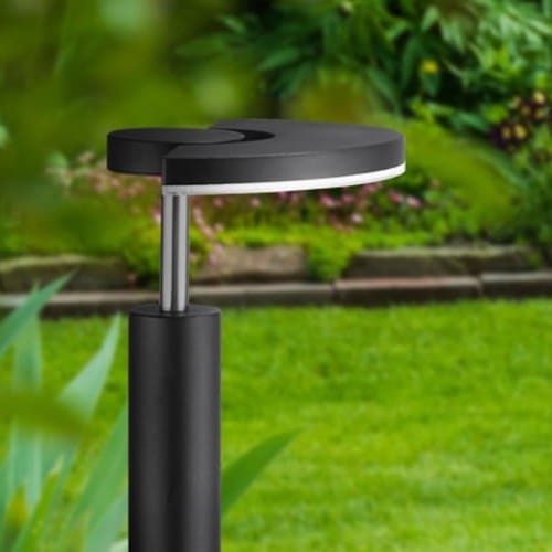 Lampa ogrodowa słupek LED MODENA o wysokości 54 cm, czarna, z okrągłą górną częścią i aluminiowym wykończeniem. Umieszczona w zielonym otoczeniu, z delikatnym tłem ogrodu i kwiatów.