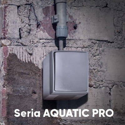 Gniazdo natynkowe AQUATIC IP44 w szarym kolorze, umieszczone na betonowej ścianie. Posiada zaokrąglone krawędzie, czytelne logo serii AQUATIC PRO na dolnej części zdjęcia.