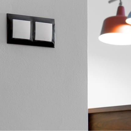 Łącznik krzyżowy podwójny Simon 54 w kolorze białym, mocowany na ścianie. Produkt o prostokątnym kształcie z dwoma przyciskami oraz minimalistycznym designie, umieszczony w jasnym otoczeniu z lampą w tle.
