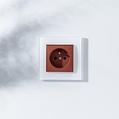Ramka pojedyncza kwadratowa Karlik ICON w kolorze białym, z efektem szkła, z widocznym gniazdem elektrycznym w kolorze brązowym. Ujęcie frontalne na białym tle, podkreślające prosty, nowoczesny design.