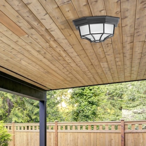 Czarna lampa sufitowa zewnętrzna MILANO Rabalux 8346, umieszczona na drewnianym suficie, z przezroczystym kloszem w kształcie sześciokąta, z widocznymi detalami metalowej ramy. Tło z ogrodzeniem i zielenią.