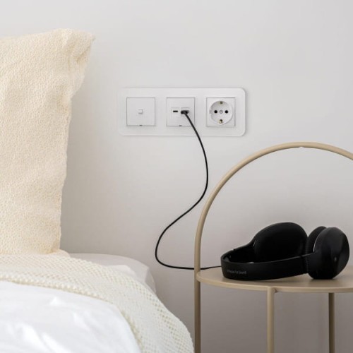 Łącznik pojedynczy Karlik ICON w białym kolorze, w stylu amerykańskim, zamontowany na ścianie obok łóżka. Widoczna wtyczka USB oraz gniazdo elektryczne, obok słuchawki na metalowej podstawce. Tło w jasnym kolorze, z poduszką i narzutą.