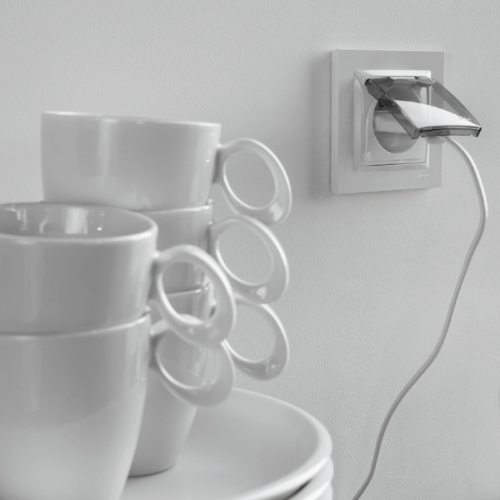 Gniazdo hermetyczne Simon 10 z uziemieniem, z klapką dymną, w kolorze białym. Zainstalowane na ścianie, z widocznym kablem elektrycznym. W tle stos białych filiżanek z uchwytami.