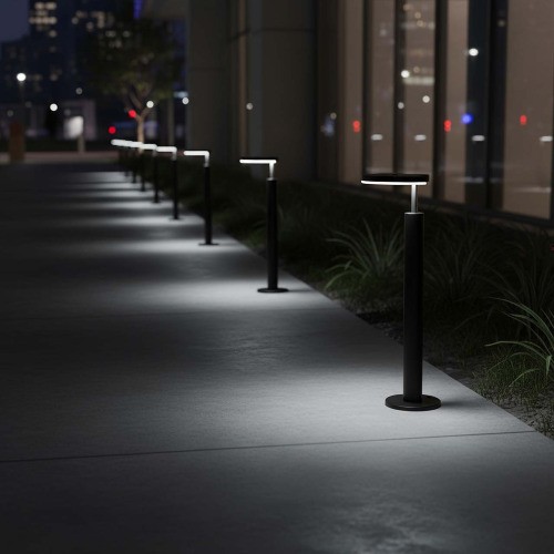 Lampa ogrodowa słupek LED MODENA o wysokości 54 cm w kolorze czarnym. Produkt stoi na okrągłej podstawie, z prostokątnym źródłem światła na górze. Widoczna linia lamp w oświetlonym otoczeniu, na tle nawierzchni i roślinności.