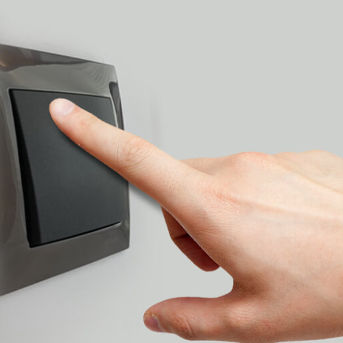 Moduł sterujący Wi-Fi ActionBox Blebox 012640 w kolorze czarnym, umieszczony na ścianie. Widoczna ręka naciskająca na przycisk, co sugeruje aktywację urządzenia. Tło jasnoszare, minimalistyczne.