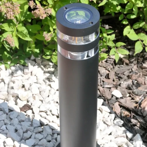 Lampa ogrodowa słupek RINO o wysokości 44 cm, okrągła, w kolorze czarnym. Wykonana z metalu, z przezroczystym elementem u góry, znajduje się na tle kamieni i zieleni. Stoi w pozycji pionowej, z widocznym oprawą E27.