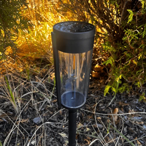 Słupek solarny PATH o wysokości 65 cm, czarny, z przezroczystą osłoną. Ujęcie z boku, stojący w ogrodzie na tle roślin. Wbudowany panel słoneczny na górze, metalowy korpus i przezroczysta dolna część, pozwalająca na emitowanie światła.