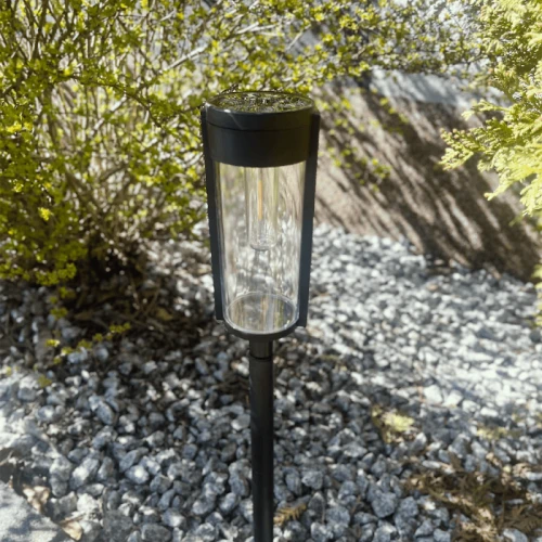 Słupek solarny o wysokości 65 cm, w kolorze czarnym, z przezroczystym kloszem. Ustawiony pionowo na podłożu, z widocznym panelem słonecznym na górze. Otoczony drobnymi kamieniami i zielonymi krzewami, idealny do oświetlenia ścieżek. Zestaw dwupak.