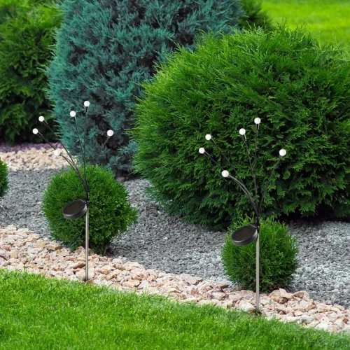 Lampa solarna Fiorini z sześcioma diodami LED, o wysokości 70 cm, z czarną podstawą i białymi kulkami na smukłych ramionach. Umieszczona w ogrodzie, wśród zielonych krzewów i kamieni dekoracyjnych.
