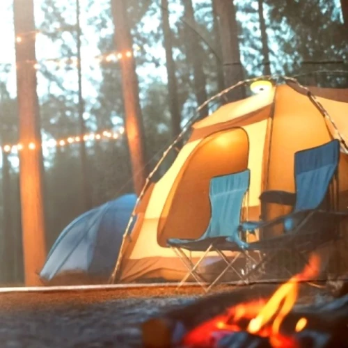Lampka campingowa USB 1W z girlandą świetlną 8m, umieszczona w lesie, przy namiocie w kolorze beżowym. Widoczne dwa niebieskie krzesła campingowe oraz ognisko z płomieniem. W tle widać girlandę świetlną, tworzącą przytulną atmosferę.