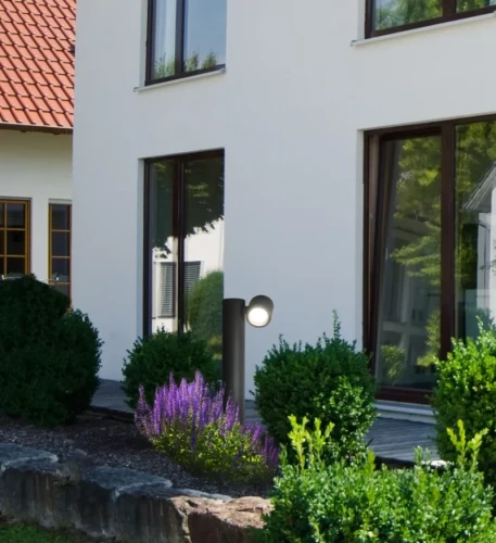 Lampa ogrodowa stojąca słupek PINO GU10 o wysokości 44 cm w kolorze czarnym, umieszczona w ogrodzie obok budynku, z widocznym źródłem światła i otoczeniem roślinności, w tym niskimi krzewami i kwiatami.
