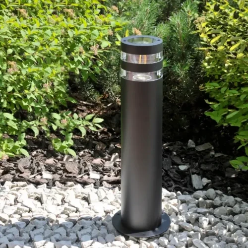 Lampa ogrodowa słupek RINO o wysokości 44 cm, okrągła, czarna, z przezroczystymi paskami na górze. Stoi na podłożu z drobnych kamieni, w otoczeniu zieleni. Posiada miejsce na żarówkę E27.