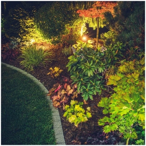 Reflektor ogrodowy LED BLAKE 5W IP65 w kolorze czarnym, emitujący światło o temperaturze barwowej 4000K, oświetla roślinność w ogrodzie, umieszczony wśród zieleni i kwiatów, tworząc przyjemną atmosferę nocnego krajobrazu.