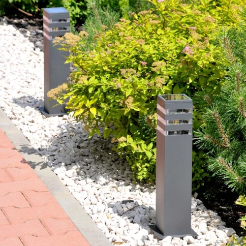 Słupek ogrodowy TEKSAS E27 o wysokości 44 cm w kolorze grafitowym, prostokątny kształt z metalowym wykończeniem i dekoracyjnymi szczelinami u góry, umieszczony w zielonym otoczeniu na tle kamieni.