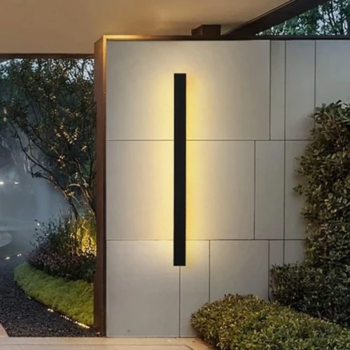 Oprawa elewacyjna LED LARA 45W o długości 150 cm, w kolorze czarnym, zamontowana na jasnej ścianie. Emituje ciepłe światło, tworząc efektowne oświetlenie. W tle widoczna zieleń oraz elementy architektury krajobrazu.
