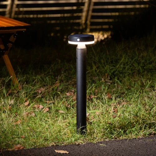 Lampa solarna z czujnikiem ruchu Gwinea, 4,5W, wysokość 50 cm. Okrągła, czarna podstawa z białym źródłem światła, umieszczona na trawie, w tle drewniany stół. Oświetlenie o barwie 4000K, działająca na energię słoneczną.