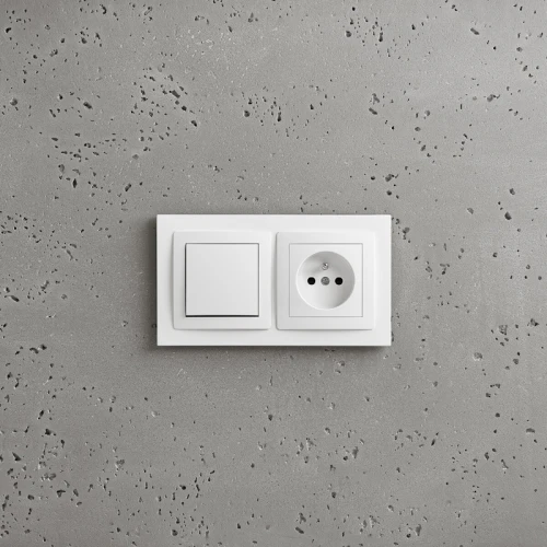 Ramka podwójna Karlik Deco w kolorze białym, montowana na ścianie, z włącznikiem po lewej stronie oraz gniazdem elektrycznym po prawej. Prosty, kwadratowy kształt, z widocznymi otworami gniazda. Tło w odcieniu szaro-białym, tekstura betonu.