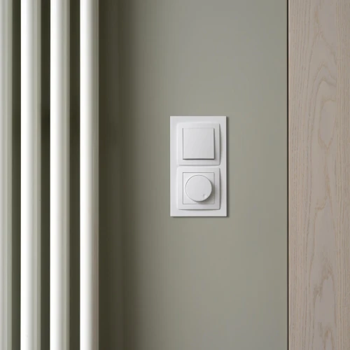 Podwójna ramka Karlik Deco w kolorze białym, montowana na ścianie. Zawiera kwadratowy przycisk i okrągły pokrętło, umieszczona obok białych rur grzewczych. Tło w odcieniu zielonym, z drewnianym elementem po prawej stronie.
