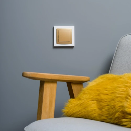 Pojedyncza ramka Karlik Deco w kolorze białym z metaliczną, złotą wkładką, zamontowana na szarej ścianie. W tle widoczny drewniany fotel z żółtą poduszką. Ramka ma prostokątny kształt i subtelne kontury.