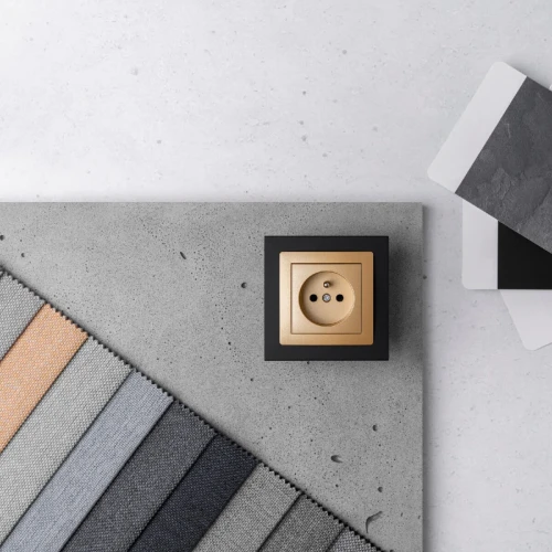 Ramka pojedyncza Karlik Deco w kolorze czarnym matowym z metalową osłoną w kolorze złotym. Umieszczona na szarej powierzchni betonu, obok próbek materiałów w różnych odcieniach. Widok z góry, produkt w pozycji stojącej.