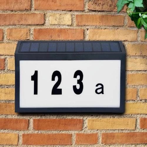 Kinkiet elewacyjny solarny LED z numerem domu, czarna ramka z białą tablicą, widoczne cyfry 123 a. U góry panel słoneczny, zamontowany na ceglanej ścianie. Prosty design, odpowiedni do oznaczania adresu.