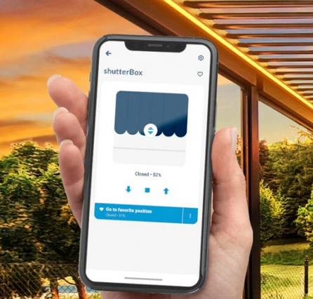 Sterownik do rolet ShutterBox v2 230V Wi-Fi na telefonie. Widoczny interfejs aplikacji z ikoną rolet, wskazującą pozycję zamknięcia na poziomie 52%. Tło zewnętrzne z zielonymi drzewami i zachodem słońca.