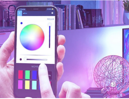 Sterownik oświetlenia RGB wLightBox Led Wi-Fi Blebox na smartfonie, z widocznym kolorowym kołem do wyboru barwy, suwakami oraz przyciskami na ekranie. W tle zabarwione na różowo oświetlenie i dekoracyjna kula.