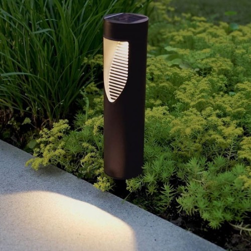 Słupek solarny ogrodowy LED, 31 cm, w kolorze czarnym. Umieszczony na tle zielonych roślin, emitujący światło. Posiada prosty, cylindryczny kształt z otworem w obudowie, który kieruje światło na powierzchnię pod spodem.
