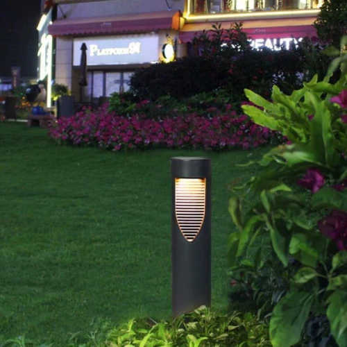 Słupek solarny ogrodowy LED o wysokości 31 cm, w kolorze ciemnoszarym, z wbudowanym oświetleniem LED. Produkt stoi pionowo na trawniku, z widocznymi paskami świetlnymi na froncie. Otoczony kwiatami i trawą, w tle widać budynek.